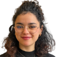 Maria Assaoud, jeune femme souriante aux cheveux bruns mi-longs, portant un haut noir ,sur fond blanc. Photo de profil professionnelle, cadrage portrait. 