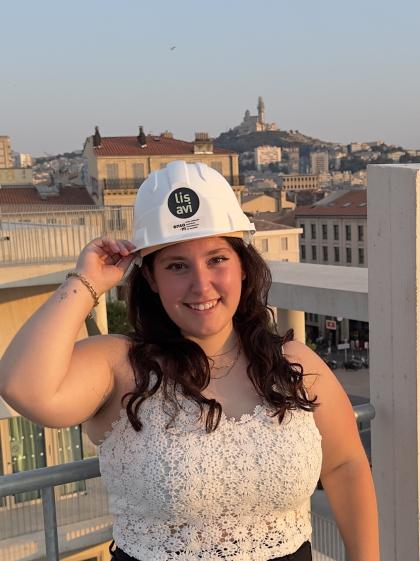 Je suis au premier plan de la photographie, avec un casque de l’IMVT sur la tête, souriante. On distingue Notre-Dame-de-la-Garde derrière.